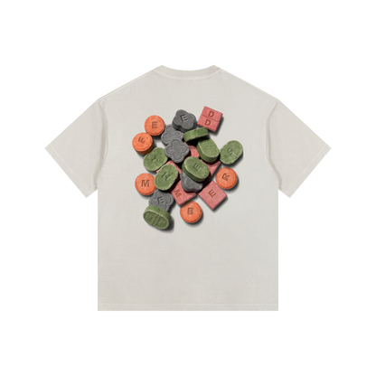 A light beige short-sleeve t-shirt with a graphic print of colorful candy-coated pills on the back, displayed against a solid white background. Front embroider Feed The Members logo in green.