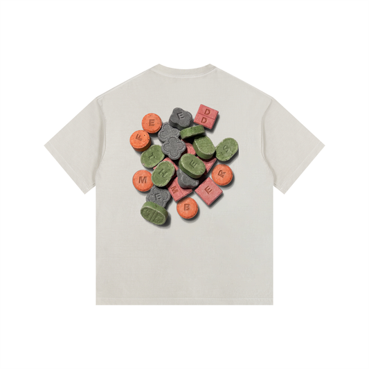 A light beige short-sleeve t-shirt with a graphic print of colorful candy-coated pills on the back, displayed against a solid white background. Front embroider Feed The Members logo in green.